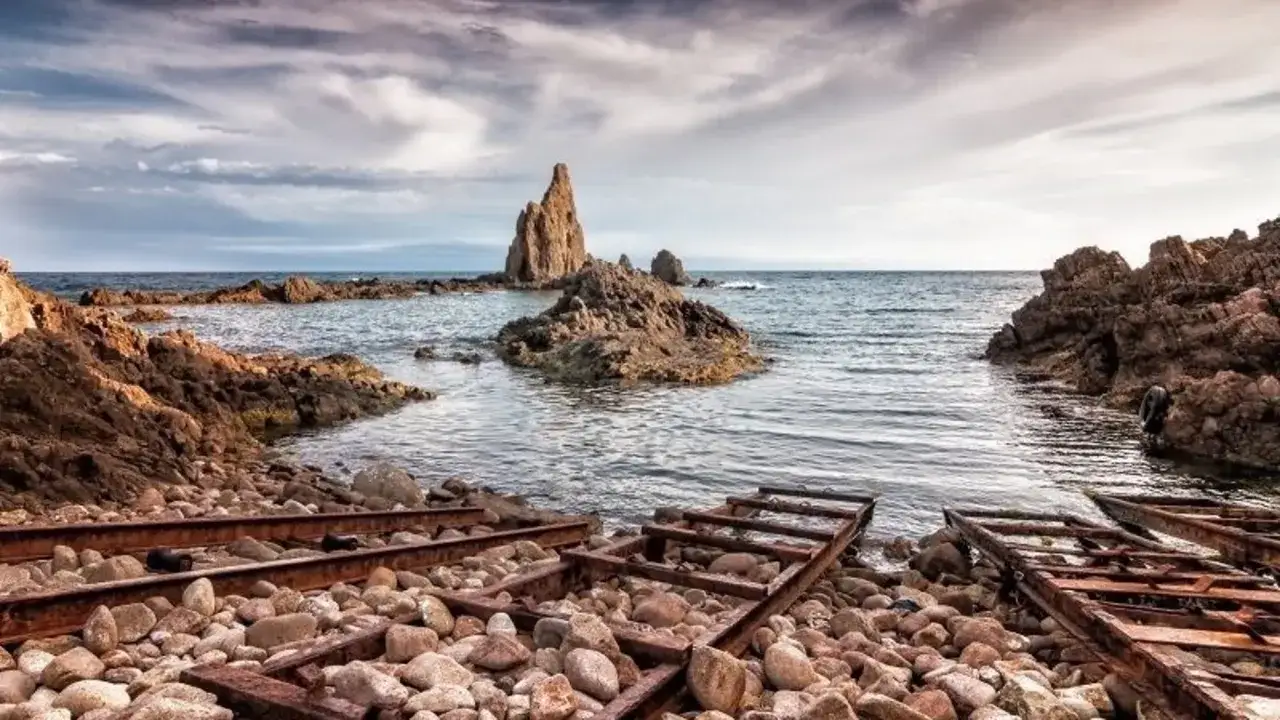 El mirador de las sirenas, con rocas escarpadas y restos de vías oxidadas, se asoma al mar bajo un cielo nublado.