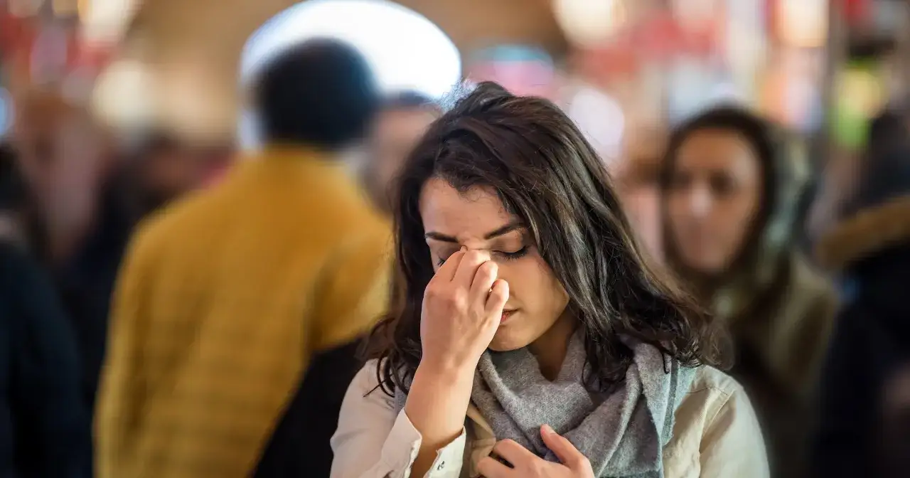 Kobieta zamyka oczy, masując czoło. Otaczają ją niewyraźne sylwetki ludzi, co może symbolizować jej fobię społeczną i poczucie przytłoczenia.