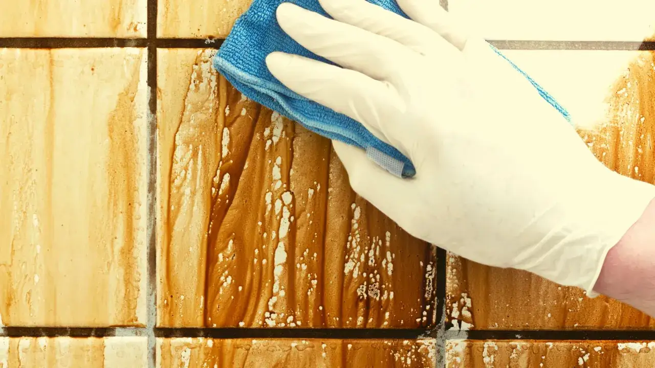 Main tenant nettoie un mur blanc jauni avec une éponge bleue. La saleté brune coule sur les carreaux.