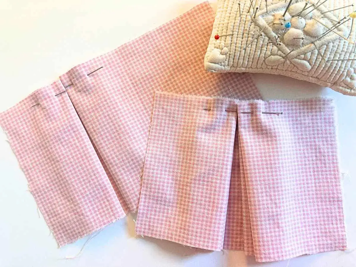 Tissu à carreaux roses avec des plis creux épinglés, prêt pour la couture. Un coussin à épingles est visible en haut à droite.