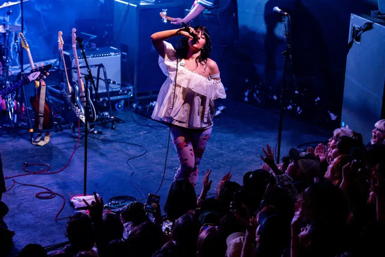 A singer belts into a mic on stage, her floral leggings a vibrant contrast to the blue stage lights. The crowd cheers, eager for the next song on the mannequin pussy setlist.