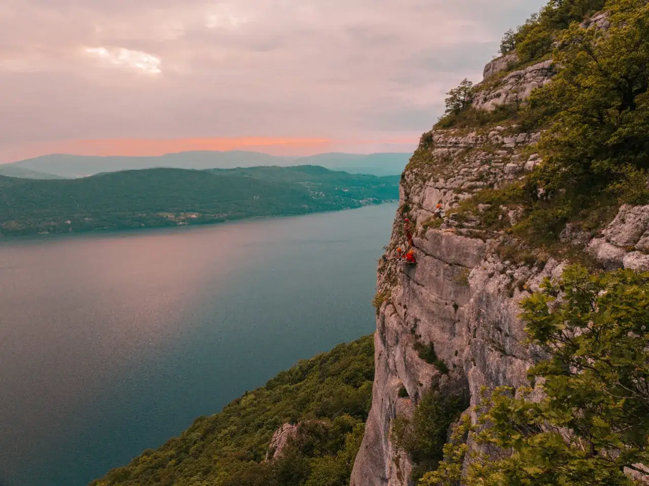 Des grimpeurs s'installent sur une **cabane falaise** surplombant un lac paisible, sous un ciel aux teintes orangées.