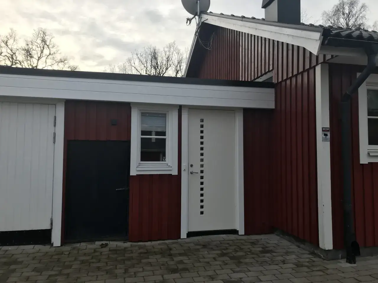 R&ouml;tt hus med vita vindskivor och takr&auml;nnor. En vit d&ouml;rr med fyrkantiga f&ouml;nster och en svart d&ouml;rr syns.