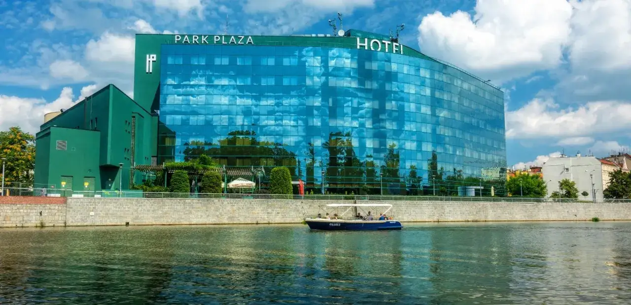 Hotel HP Plaza Wrocław z zieloną fasadą i lustrzanymi oknami odbijającymi niebo. Po rzece płynie statek wycieczkowy.