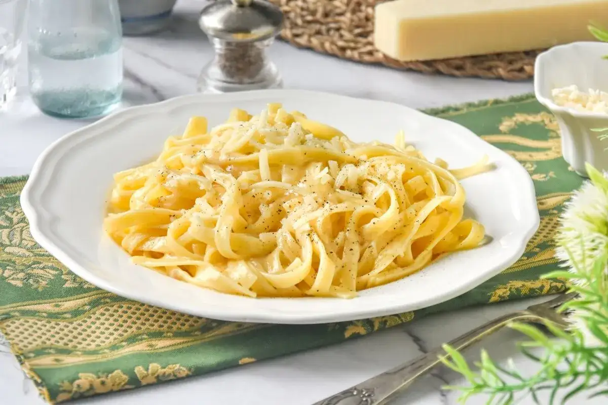 Un plato de pasta alfredo cremoso, cubierto con queso rallado y pimienta negra, servido en una vajilla blanca.