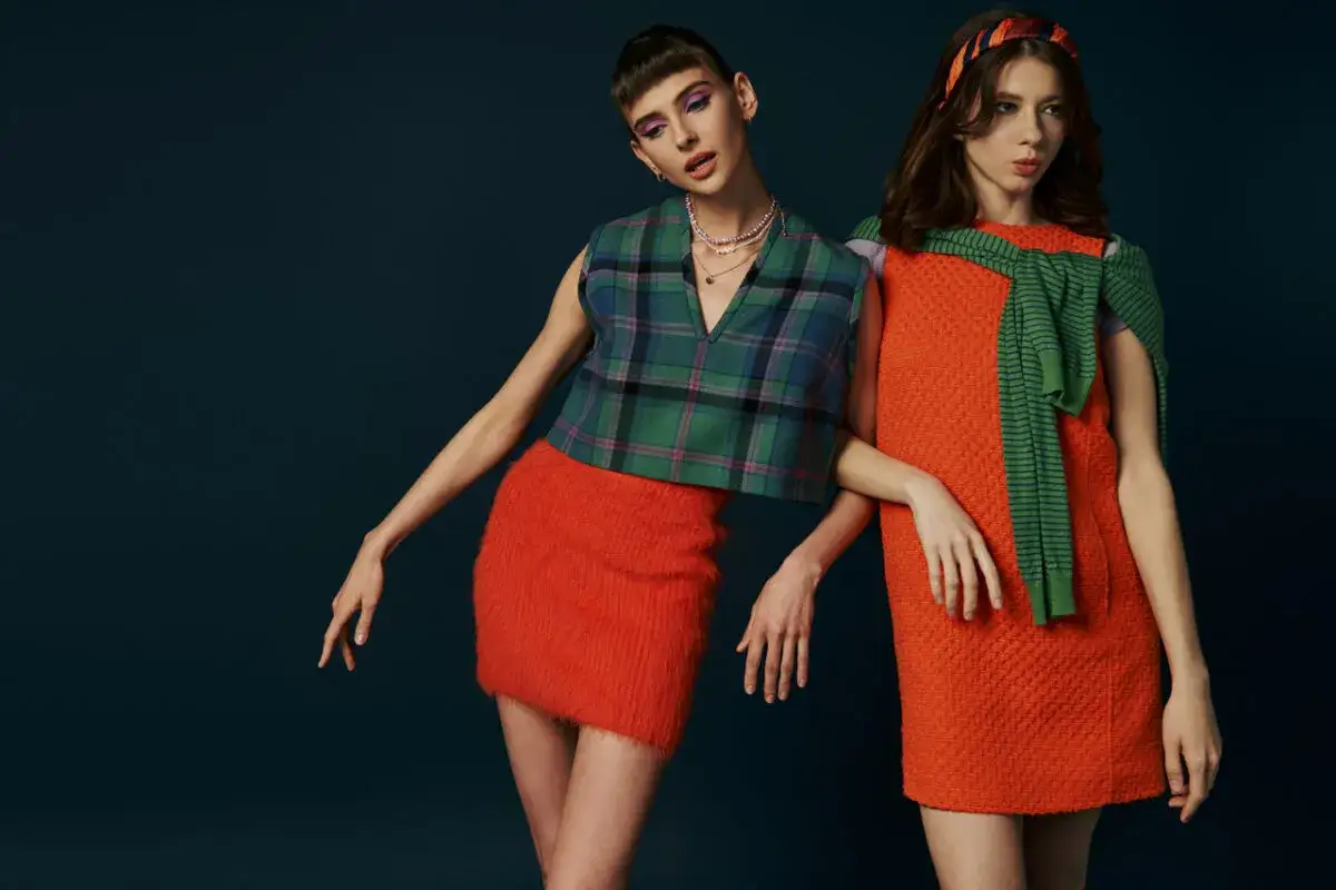 Dwie modelki w stylizacjach inspirowanych modą lat 60. Jedna w kraciastej kamizelce i pomarańczowej spódniczce, druga w pomarańczowej sukience i zielonym sweterku.
