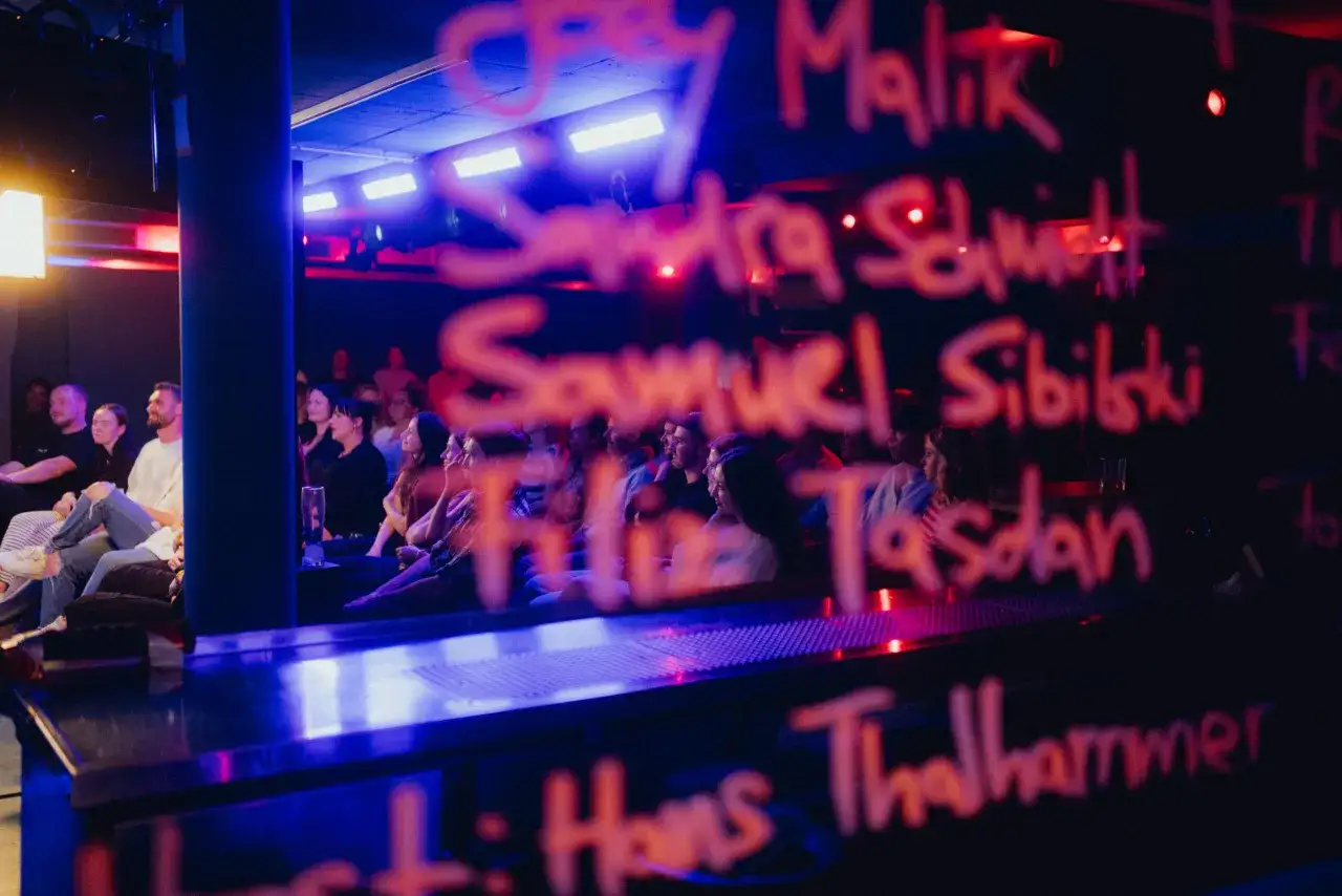 Publikum bei einer Comedy-Show in Berlin. Namen von Künstlern sind auf der Tafel geschrieben, die im Scheinwerferlicht leuchtet.