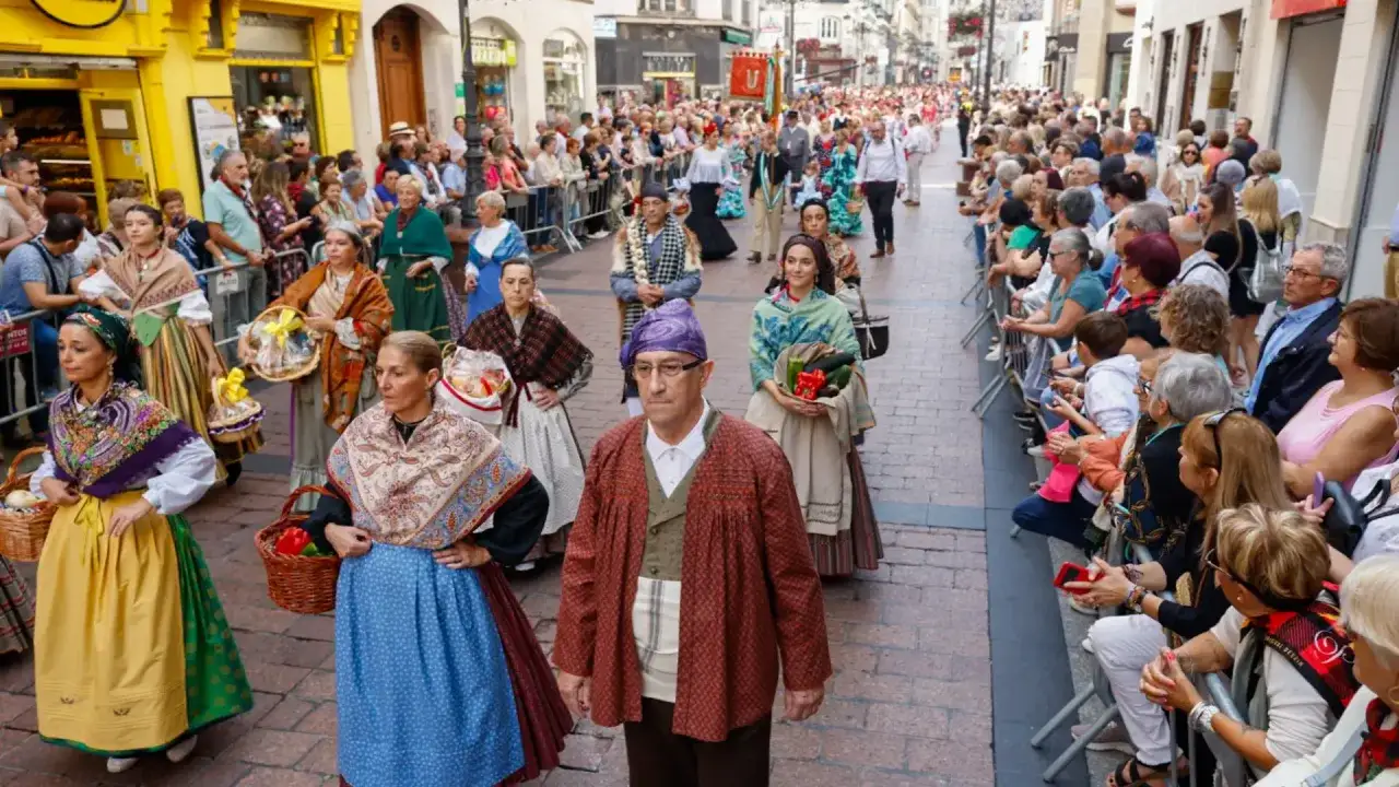 Desfile de ofrenda de frutos con trajes tradicionales. Gente observa el paso de participantes con cestas y verduras.