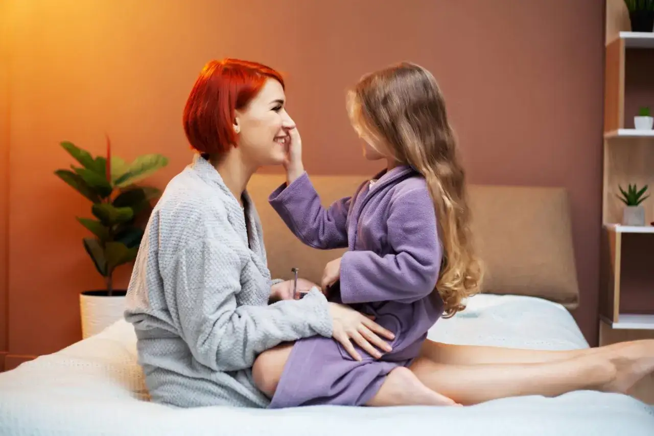 Mama i córka w szlafrokach, przytulają się na łóżku. Córka delikatnie dotyka policzka mamy. Spanie z dzieckiem 10 letnim to piękny czas.