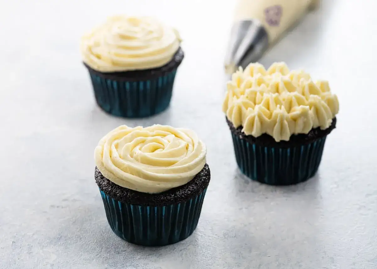 Tre mörka muffins med ljus frosting, en med rosmönster, en med små toppar. En sprits med frosting syns i bakgrunden.