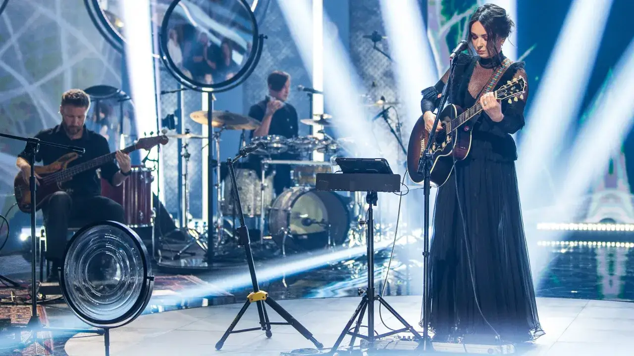 Kasia Kowalska na scenie, grając na gitarze akustycznej. W tle zespół: basista i perkusista.