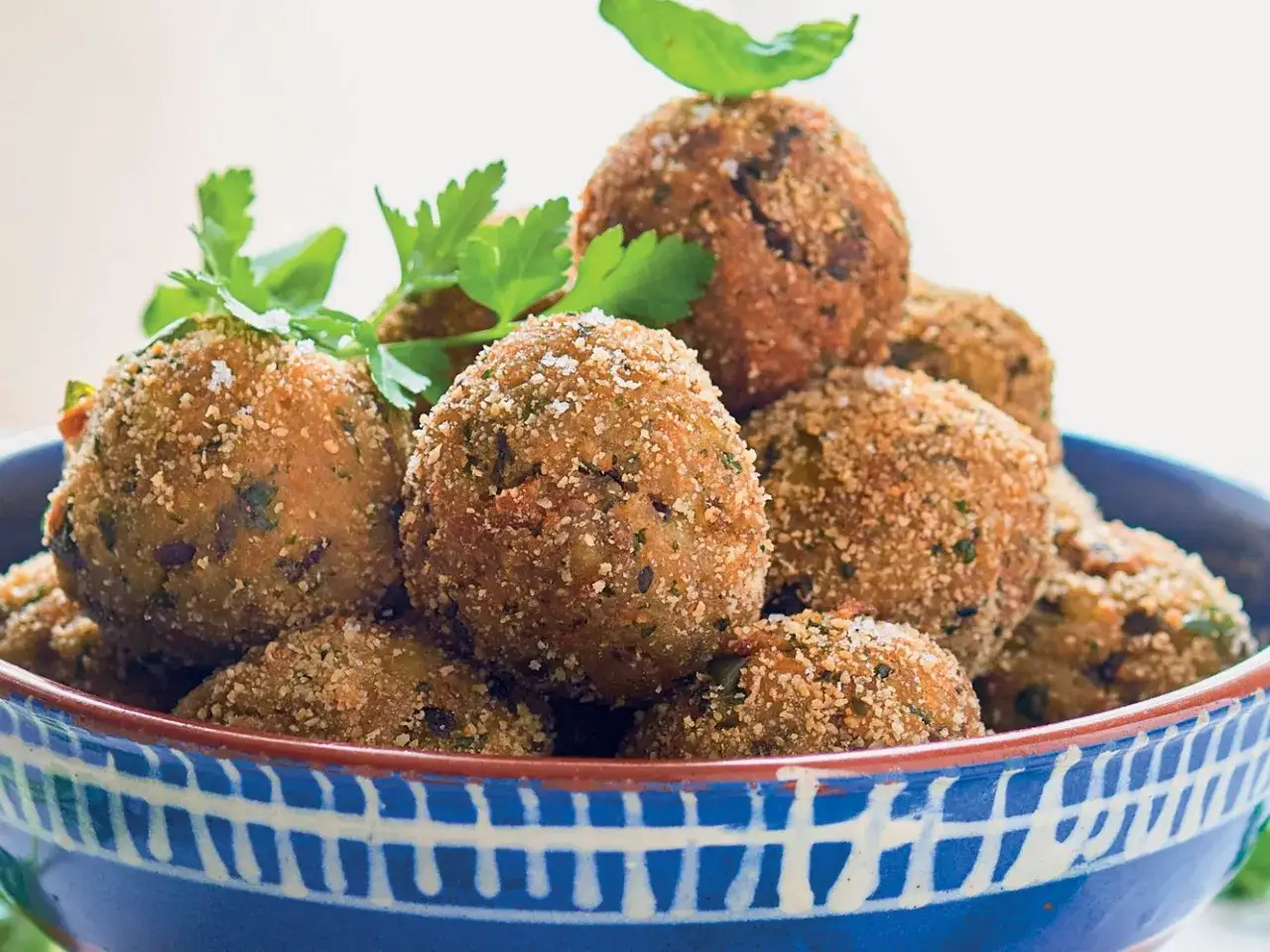 Polpette di melanzane ricetta originale, dorate e croccanti, guarnite con prezzemolo fresco e basilico, servite in una ciotola blu.