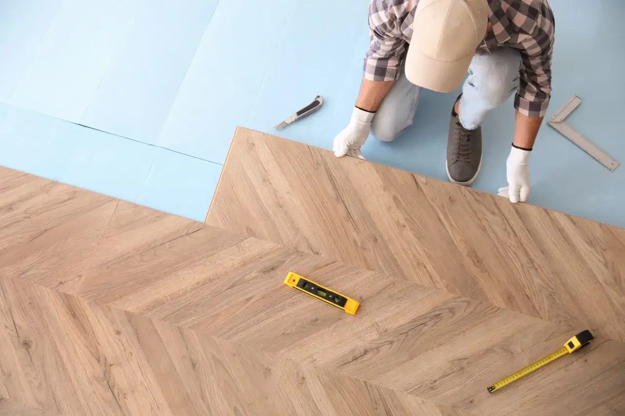 Instalator układa panele winylowe w jodełkę, pokazując, jak kłaść panele winylowe. Narzędzia takie jak poziomica i miarka są w pobliżu.