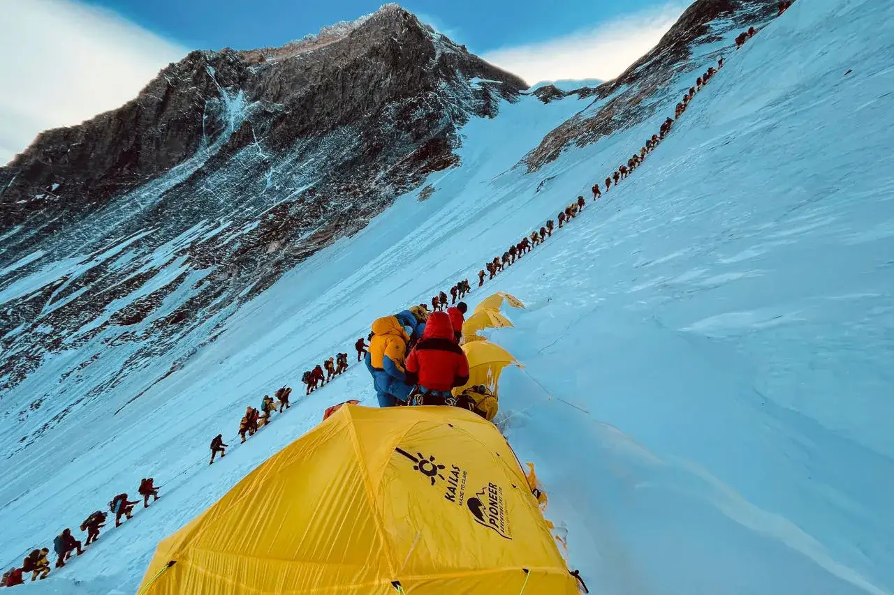 Kolejka ludzi wspina się po śnieżnym zboczu. W tle widać skaliste szczyty. Wszystko za Everest, marzenie wielu.