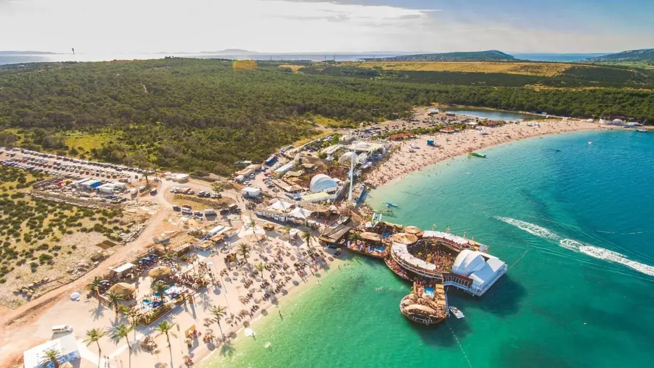 Vue aérienne d'un immense festival en Croatie, avec une plage bondée, des scènes et une structure flottante sur l'eau turquoise.