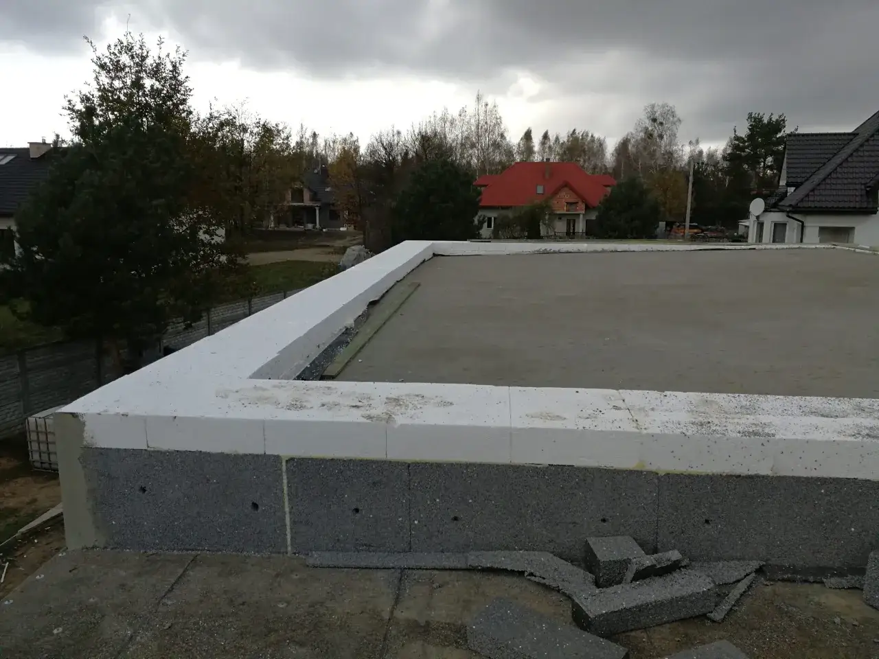 Budowa domu w toku. Widoczna warstwa izolacji z białego styropianu, czyli styropapa, otacza fundament. W tle domy i drzewa.