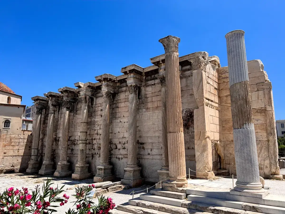 Ruinas de la Biblioteca de Adriano en Atenas, un sitio clave que ver en Atenas en 2 días. Columnas antiguas y flores rosadas bajo un cielo azul.