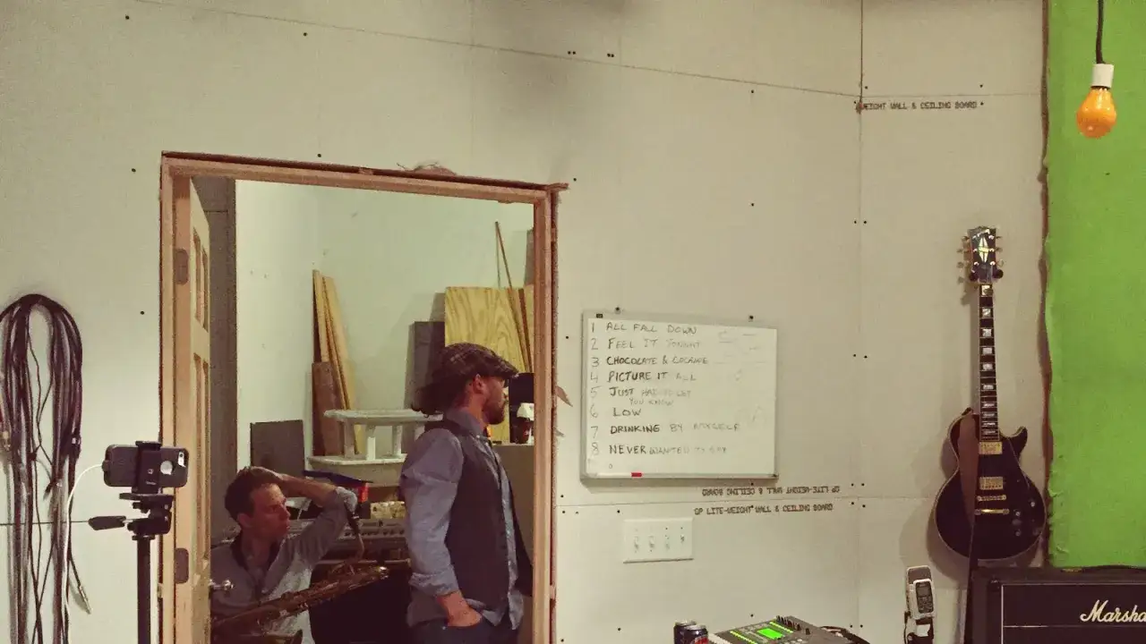 Musiker bereiten sich auf ein Musikvideo drehen mit Handy vor. Eine Setlist hängt an der Wand, eine Gitarre und ein Saxophon sind zu sehen.
