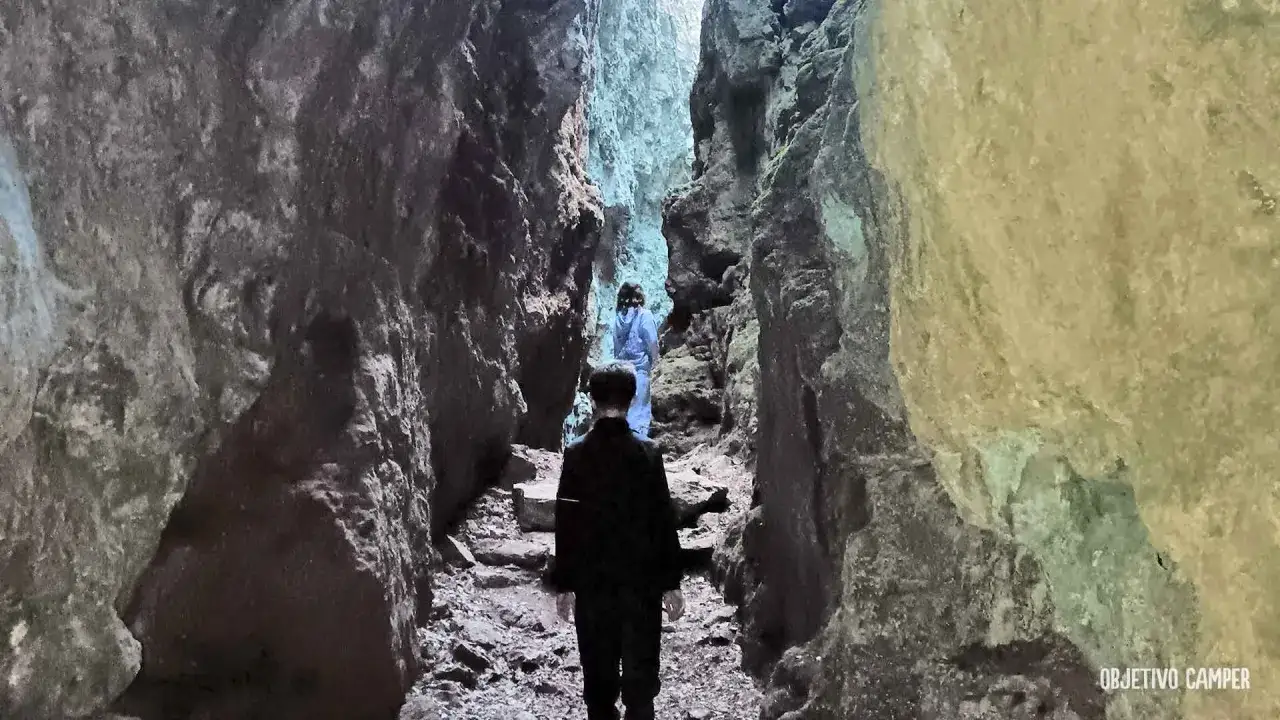 Dos personas caminan por un estrecho sendero entre imponentes paredes rocosas, adentr&aacute;ndose en los avencs de la Febr&oacute;.
