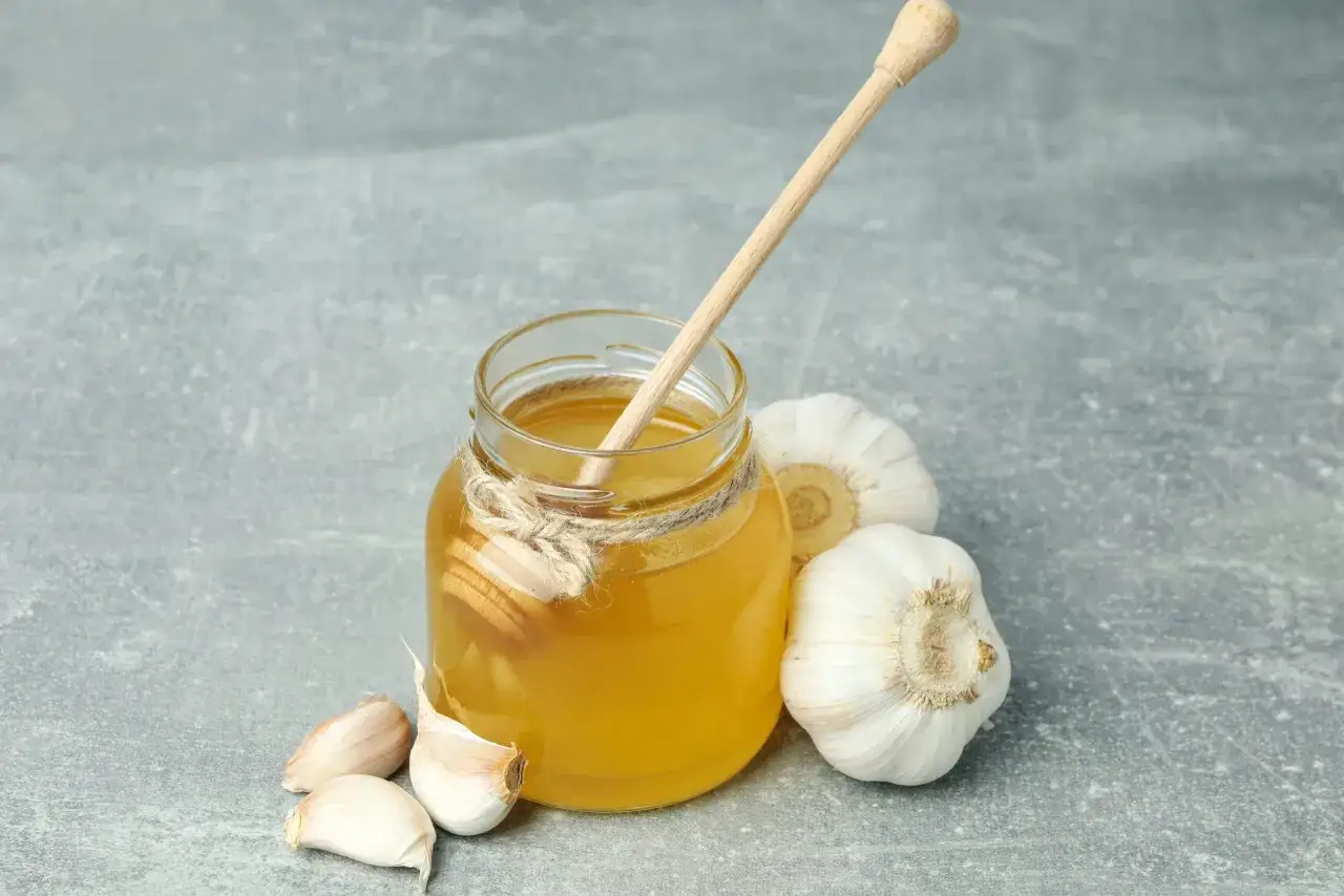 Miele dorato in un barattolo con un bastoncino, circondato da spicchi e bulbi d'aglio, un antibiotico naturale pi&ugrave; potente.