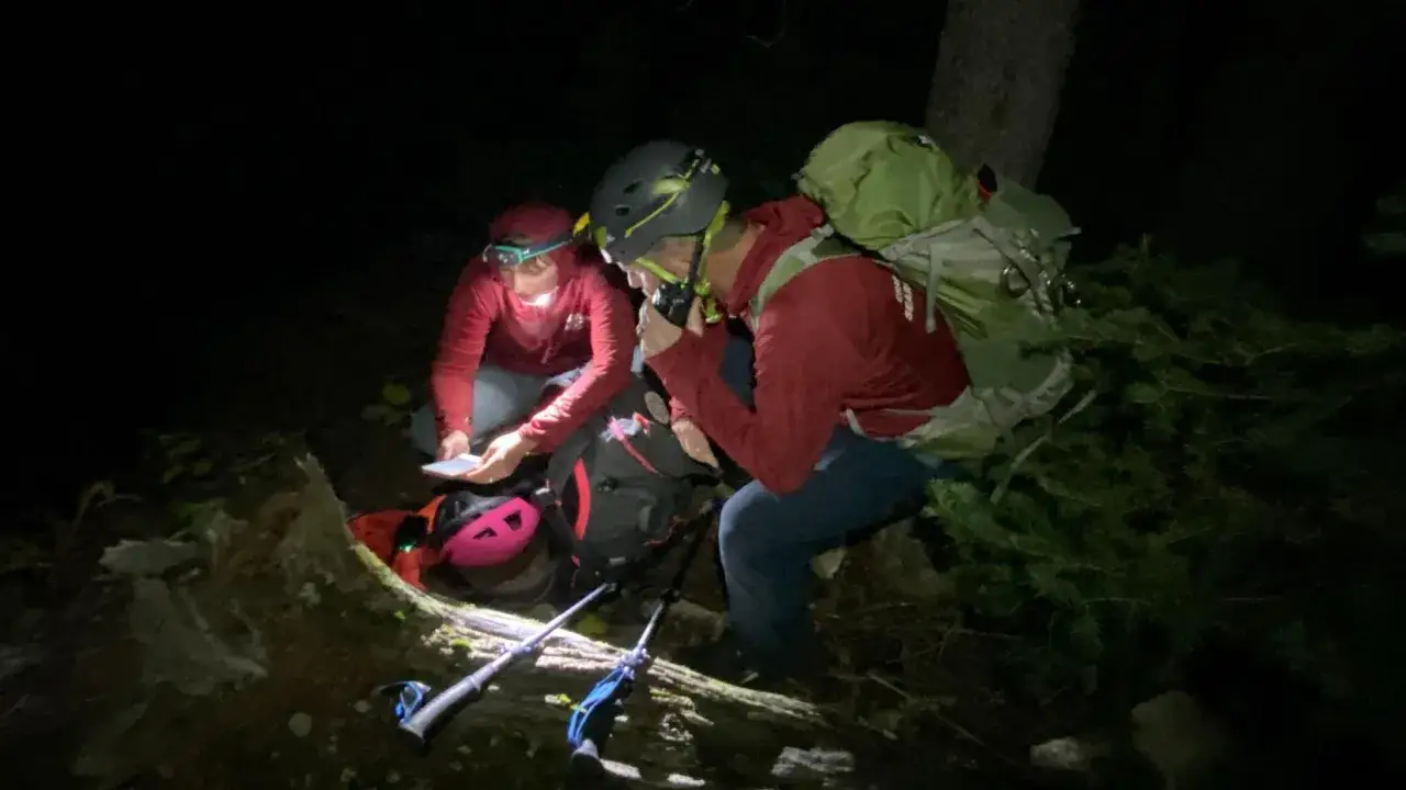 Dos excursionistas con linternas frontales y mochilas revisan un mapa en la oscuridad. Uno habla por un radio, quiz&aacute;s usando un silbato de emergencia.