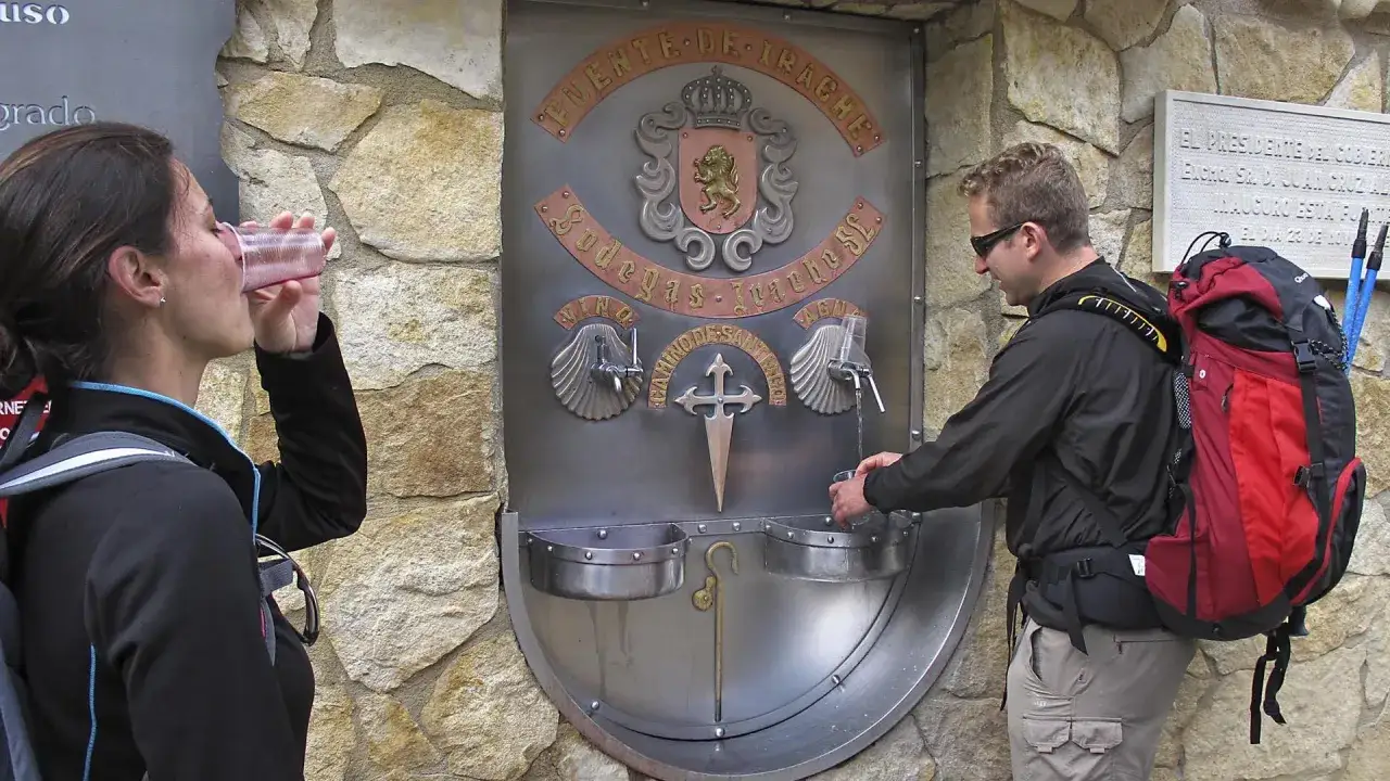 Peregrinos beben en la fuente del vino del Camino de Santiago. Un hombre llena su vaso mientras una mujer bebe.