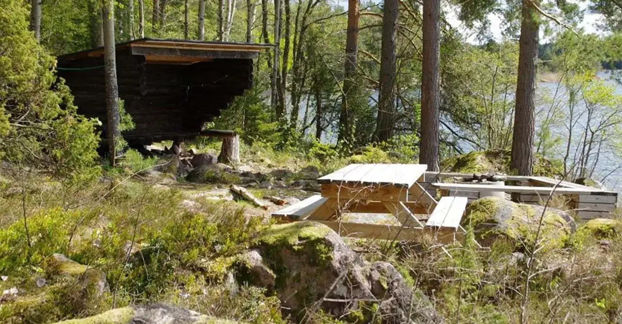 Ett rustikt vindskydd i skogen med picknickbord och grillplats vid en sj&ouml;. Perfekt f&ouml;r en paus i naturen.