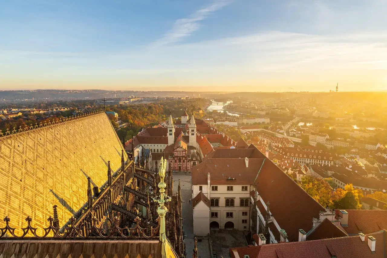 Vue panoramique du **château de Prague** baigné par le soleil levant, avec ses toits de tuiles rouges et la ville s'étendant à perte de vue.