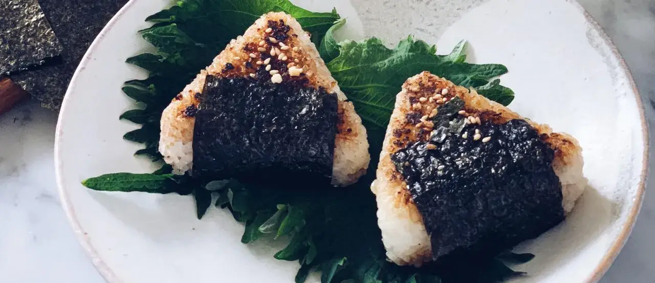 Dwa pyszne yaki onigiri, ryżowe trójkąty z nori i sezamem, podane na liściach shiso.