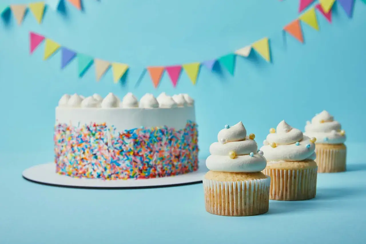 Torta bianca con codette colorate e cupcake decorati con soffice crema al burro, su sfondo azzurro con festoni.