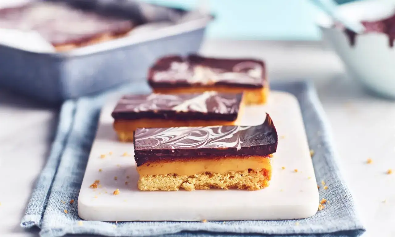 Drei saftige Millionaire Shortbread-Riegel mit Schokoladenglasur und Karamellf&uuml;llung auf einem Marmorbrett.