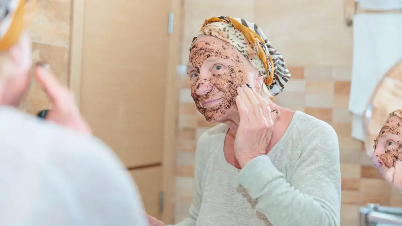 &Auml;ltere Frau mit Gesichtsmaske, die trockene Haut im Alter pflegt. Sie tr&auml;gt ein Kopftuch und schaut in den Spiegel.