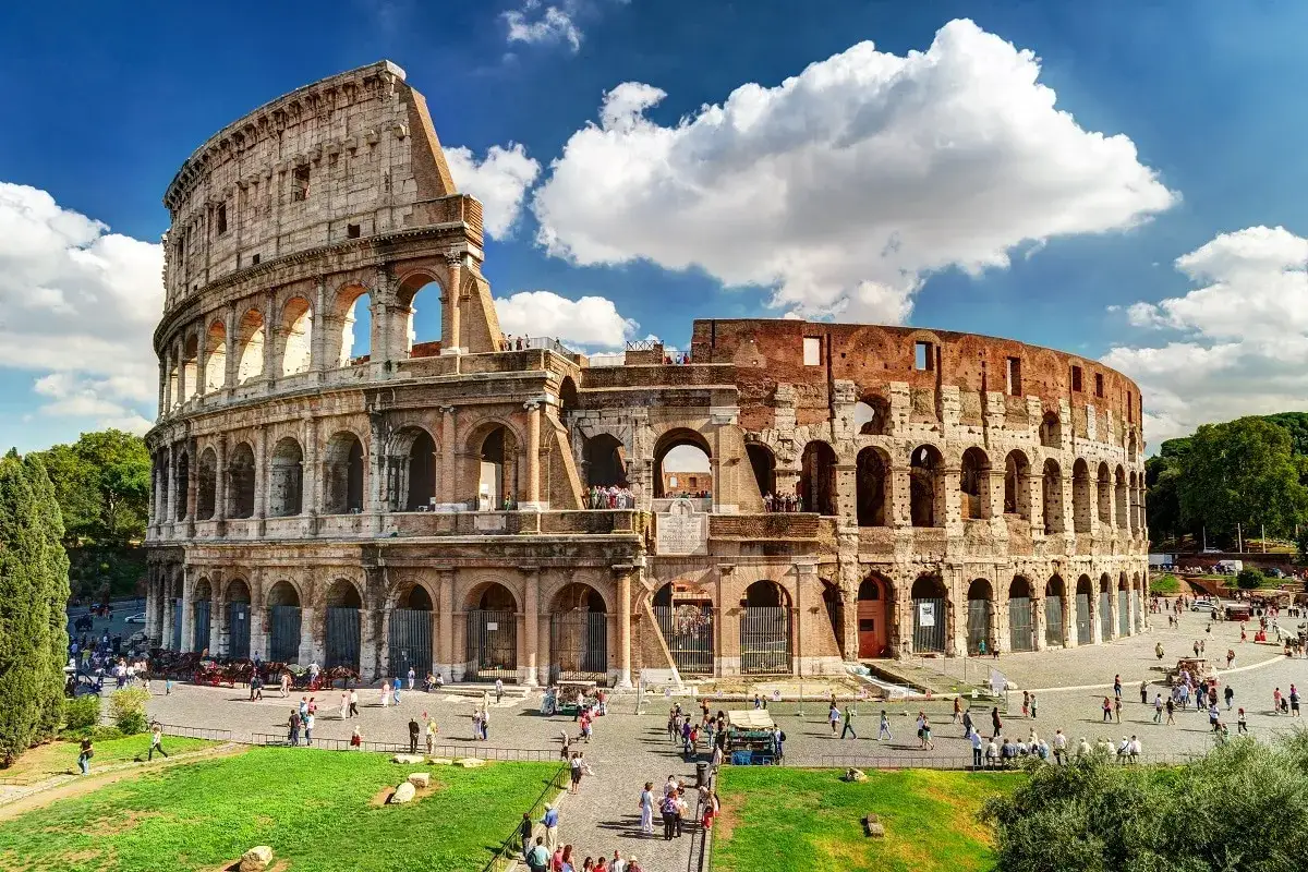 Il Colosseo, icona delle visite a Roma, si erge maestoso sotto un cielo azzurro con nuvole bianche. Turisti passeggiano nel piazzale antistante.