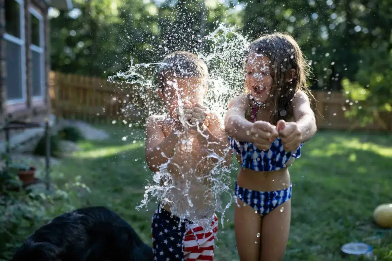 Radosne zabawy z wodą dla dzieci w ogrodzie. Chłopiec w spodenkach w amerykańską flagę i dziewczynka w bikini chlapią się wodą, tworząc fontanny.