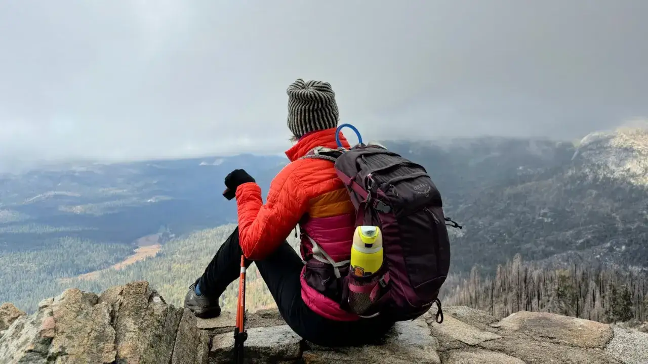 Persona con equipamiento de senderismo, mochila y bastones, sentada en la cima de una monta&ntilde;a, contemplando el paisaje nublado.