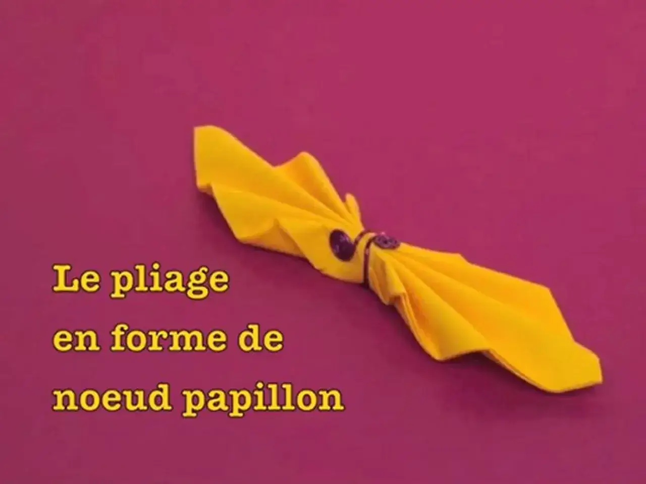 Une serviette jaune pliée en forme de noeud papillon, tenue par un anneau. Un pliage élégant pour une table festive.