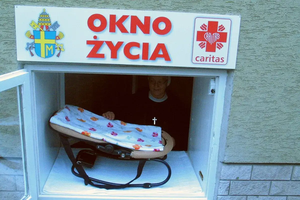 Okno życia Caritas. W środku gondola z kocykiem, obok stoi osoba w czarnym swetrze z krzyżykiem.