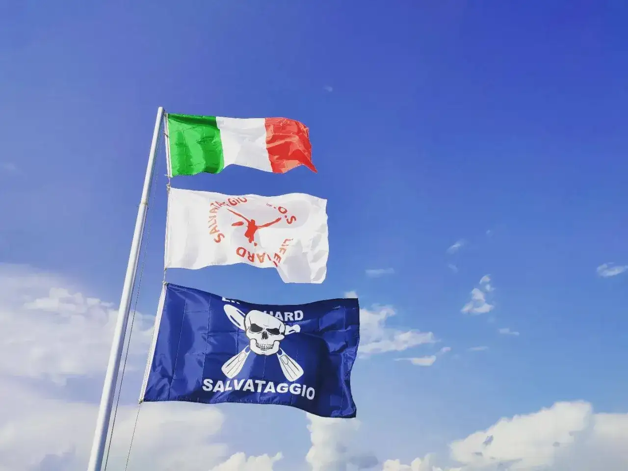Bandiera azzurra con croce bianca e teschio, bandiera bianca con salvataggio e bandiera italiana sventolano sotto un cielo azzurro.