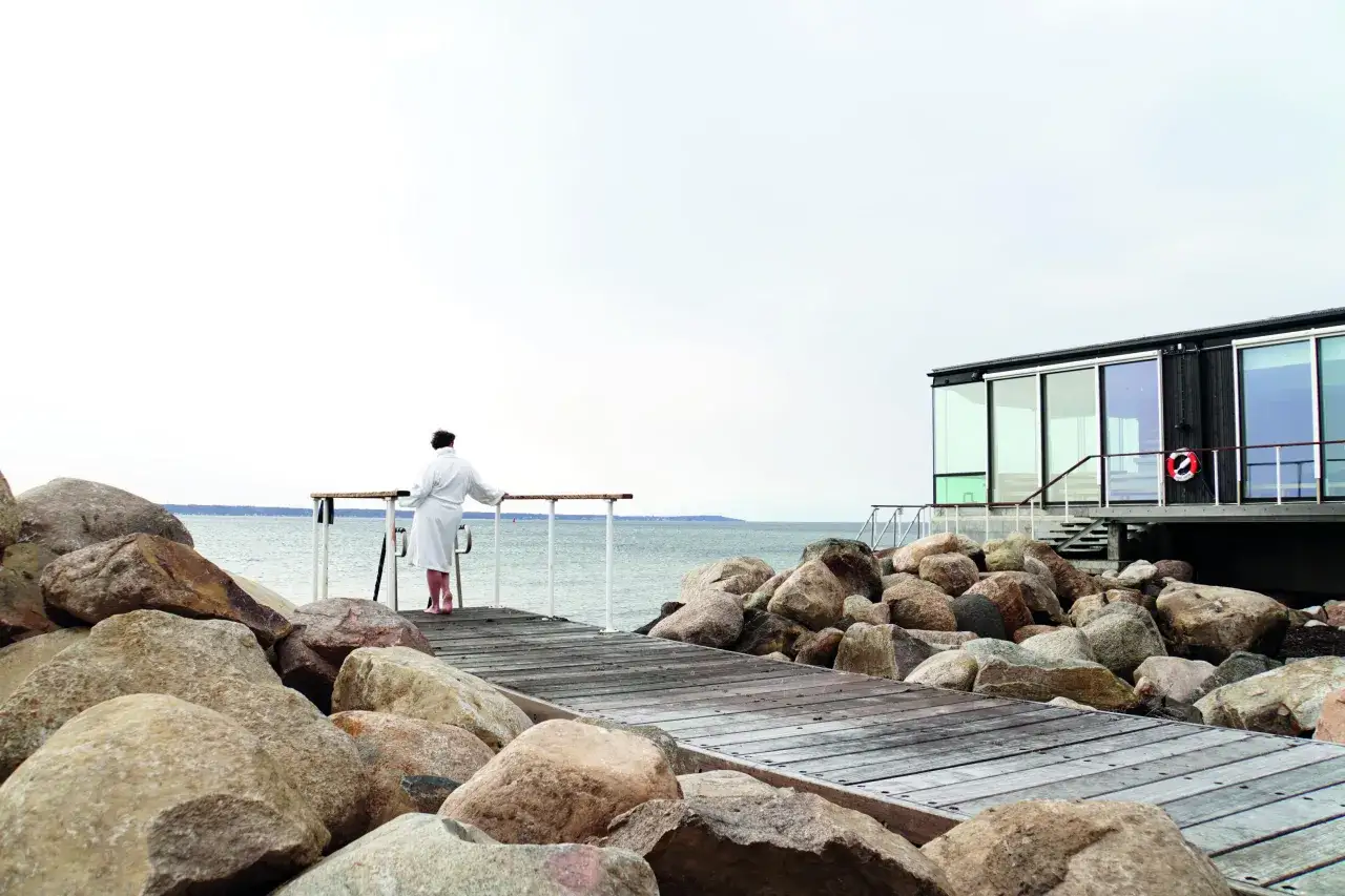En person i morgonrock njuter av utsikten vid en bastuflotte i Skåne.