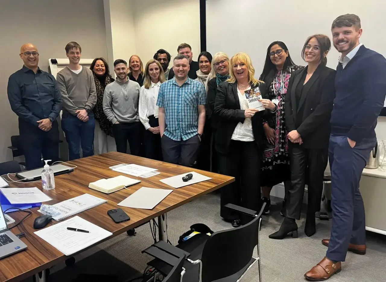 A group of professionals smiles, celebrating completion of sales leadership courses. One woman holds a book titled