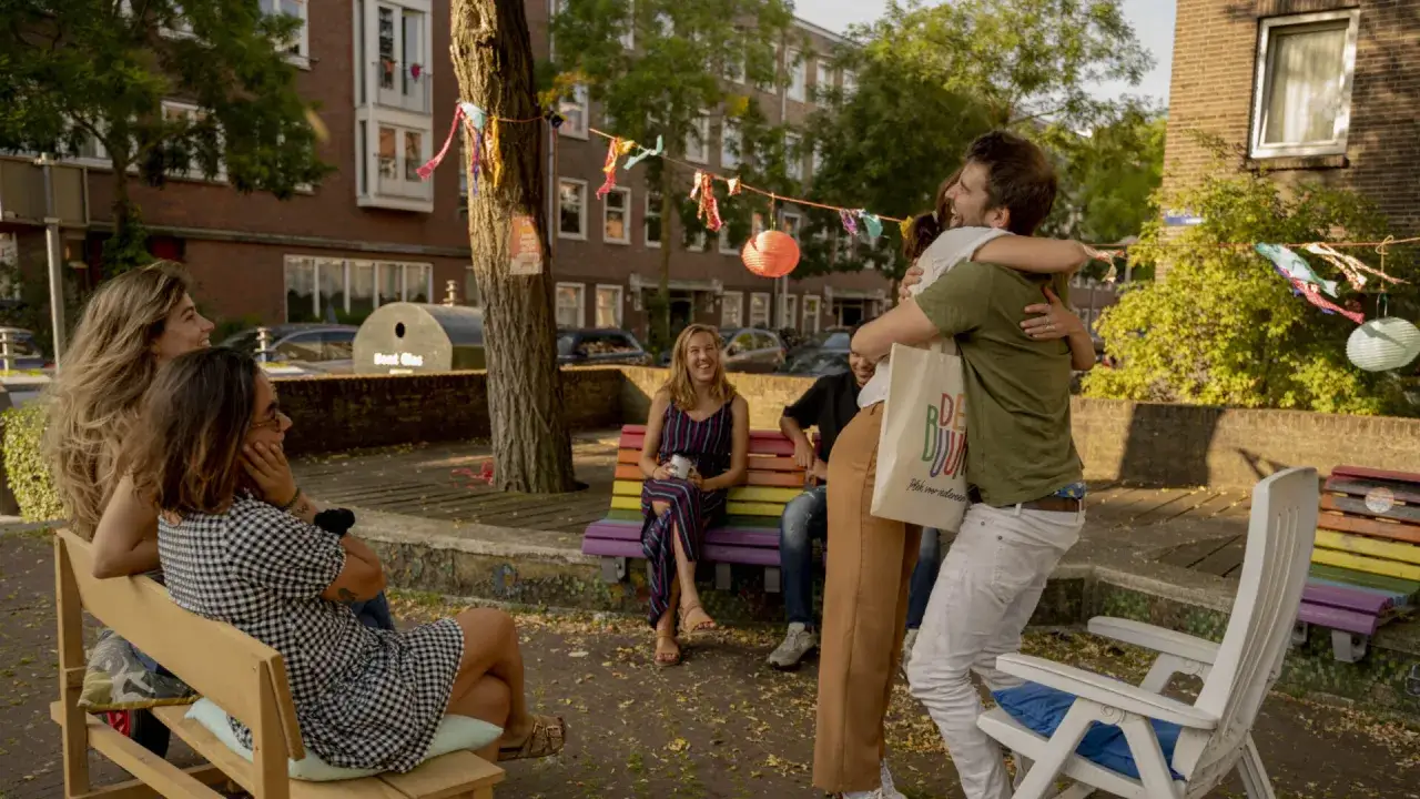 Een groep vrienden viert een hereniging in een zorgzame buurt, met vrolijke slingers en een regenboogbankje.