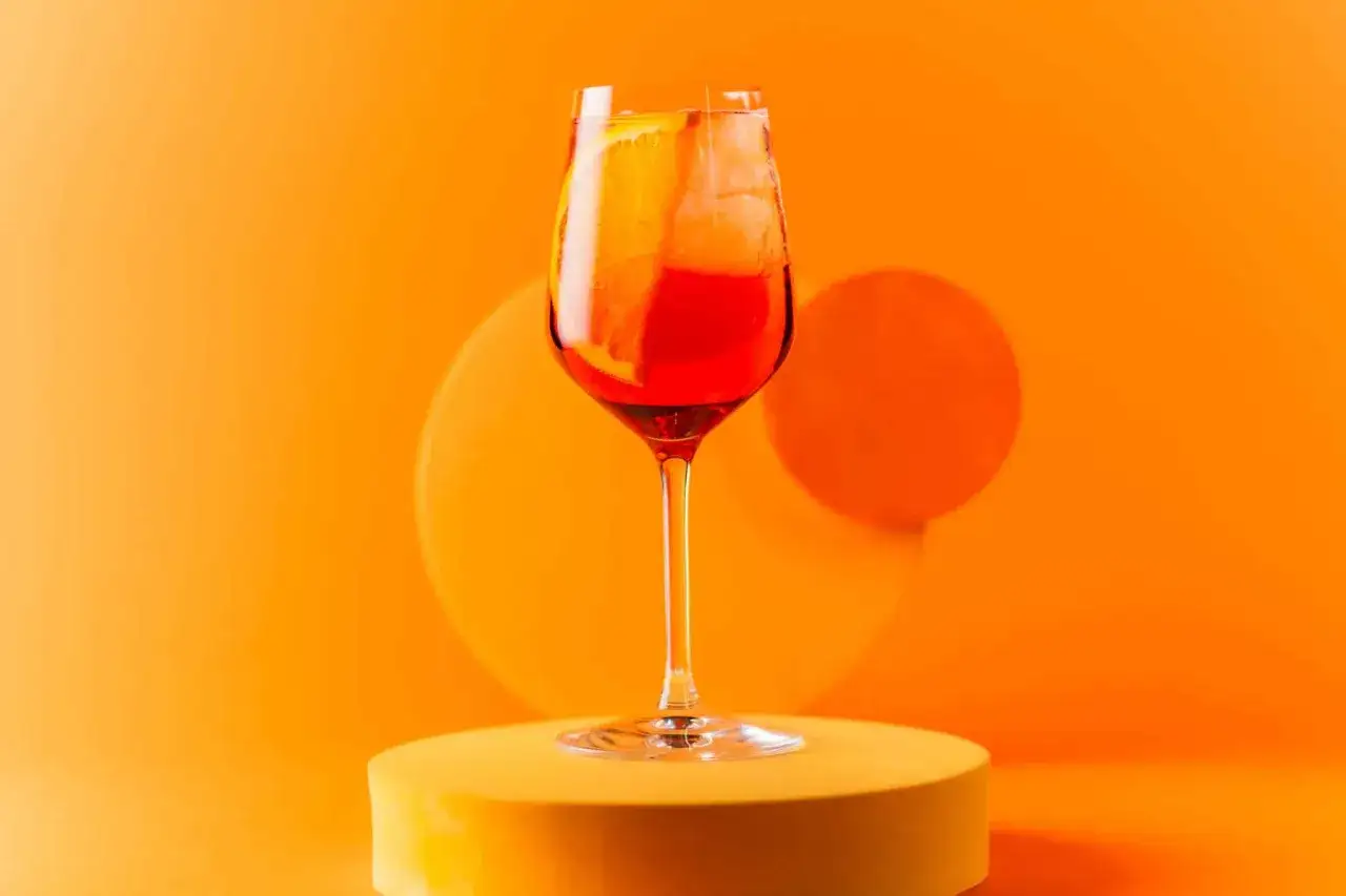 Orzeźwiający spritz campari z plasterkiem pomarańczy i lodem, prezentowany na żółtej podstawce na tle pomarańczowego gradientu.