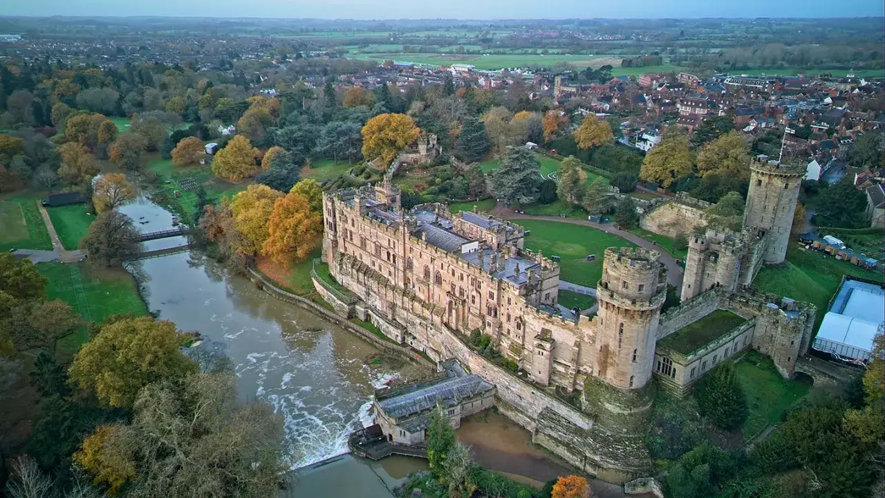 Warwick Castle, en majest&auml;tisk medeltida f&auml;stning, reser sig vid en flod omgiven av h&ouml;stf&auml;rgad natur och en stad.
