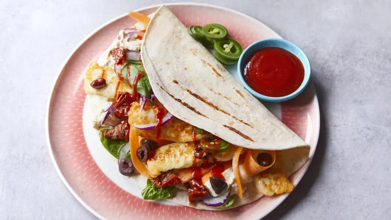 En halloumi wrap fylld med grönsaker, oliver och soltorkade tomater, serverad med en skål chilisås.