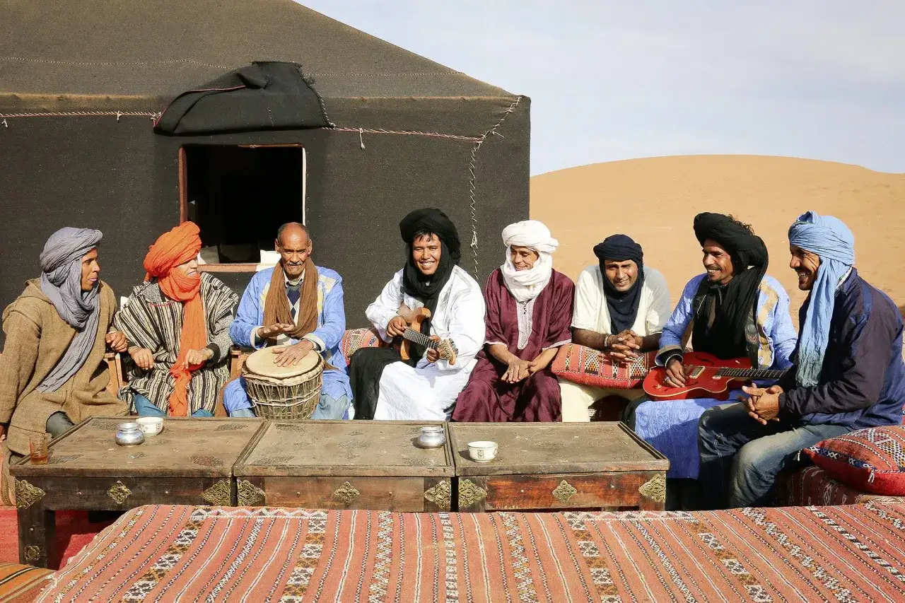 Sieben M&auml;nner in traditioneller Kleidung sitzen vor einem Zelt in der W&uuml;ste. Sie sind Marokkaner, Araber, und musizieren mit Trommel und Gitarre.