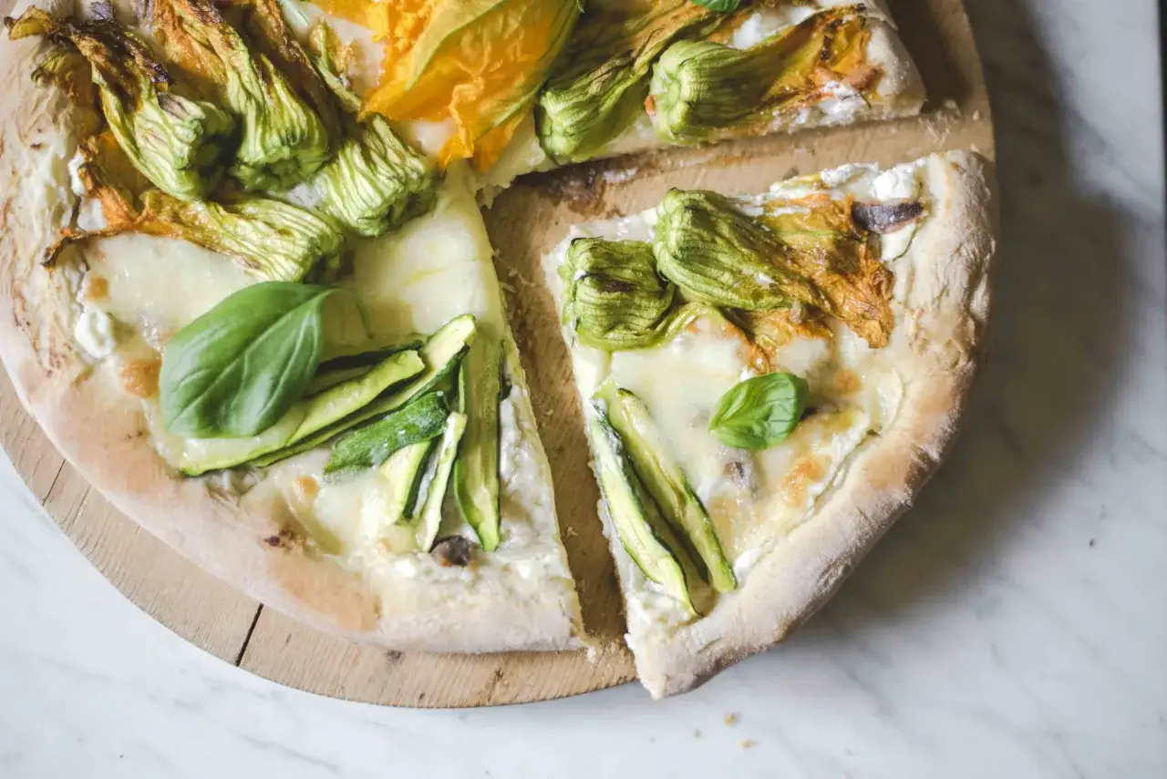 Pizza con fiori di zucca, zucchine e mozzarella filante, guarnita con basilico fresco. Un sapore estivo su un impasto croccante.