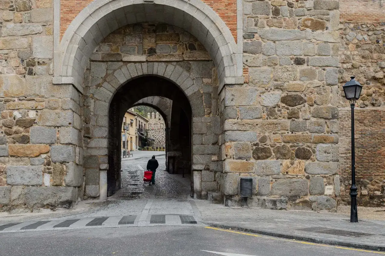 Un hombre con un carrito rojo cruza un arco de piedra en Toledo. Descubre qu&eacute; ver en Toledo y su historia.