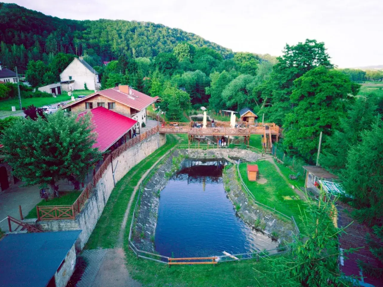 Malowniczy widok na camping w Górach Stołowych. Drewniane tarasy, restauracja z czerwonym dachem i staw otoczony zielenią.