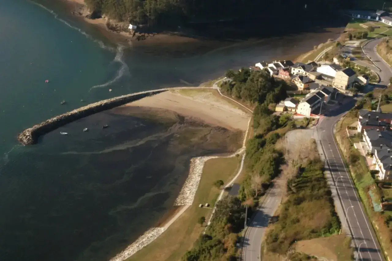 Vista aérea de la playa de los bloques Ribadeo, con un rompeolas de piedra, arena, casas y una carretera serpenteante.