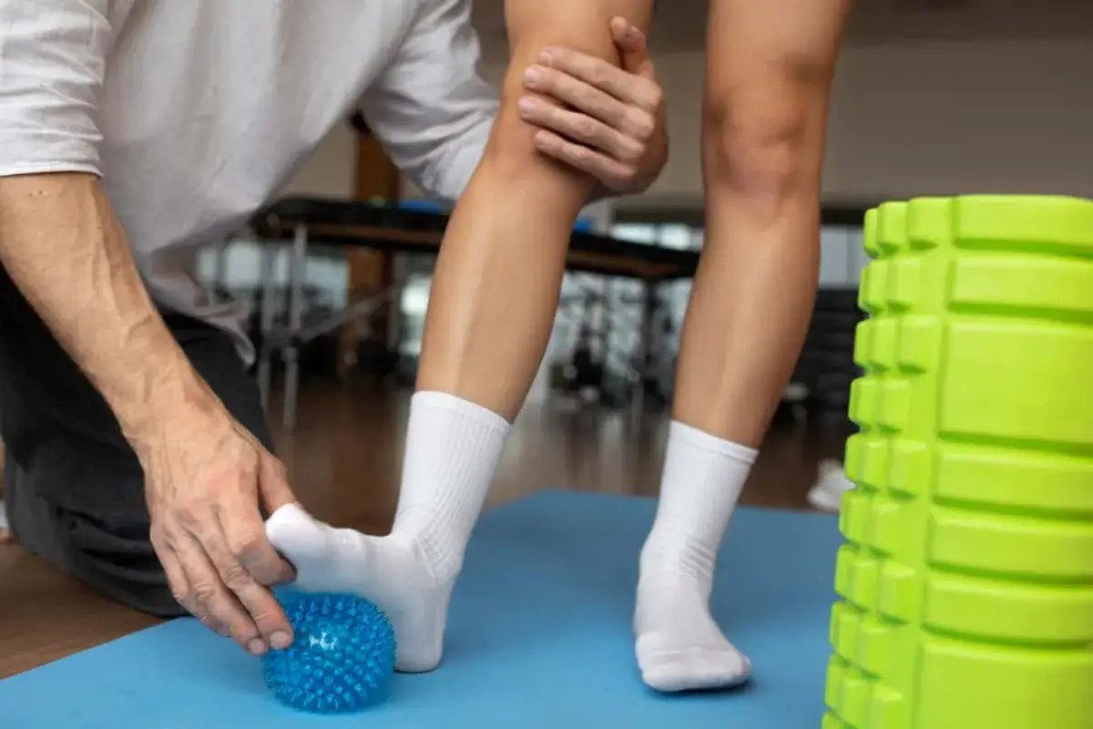 Fizjoterapeuta pomaga w domowej rehabilitacji stawu skokowego, masując stopę piłeczką. Obok zielony wałek do ćwiczeń.